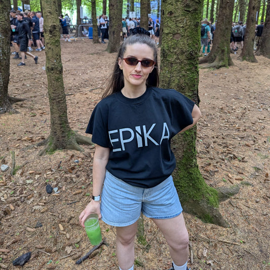 A woman standing in a forest among trees, wearing a black graphic t-shirt with the word 'EPIKA' printed in white across the chest, light blue denim shorts, and sunglasses.