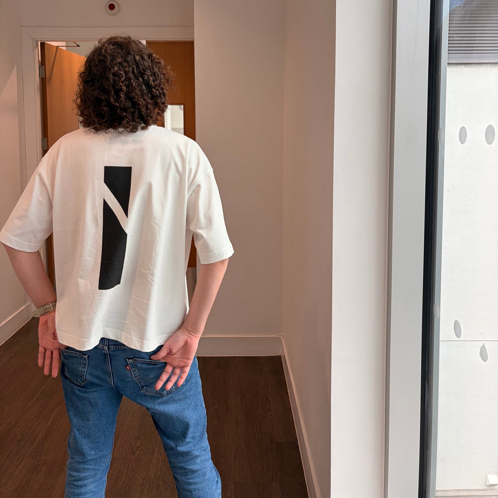 A person wearing a white short-sleeve t-shirt with a black geometric stripe detail on the back, paired with blue jeans, shown from behind in an indoor setting.