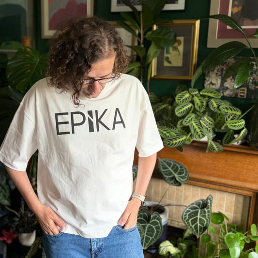 A person with curly hair and glasses wearing a white graphic t-shirt with the word 'EPiKA' printed in black across the chest, paired with blue jeans, standing in a room filled with green houseplants.