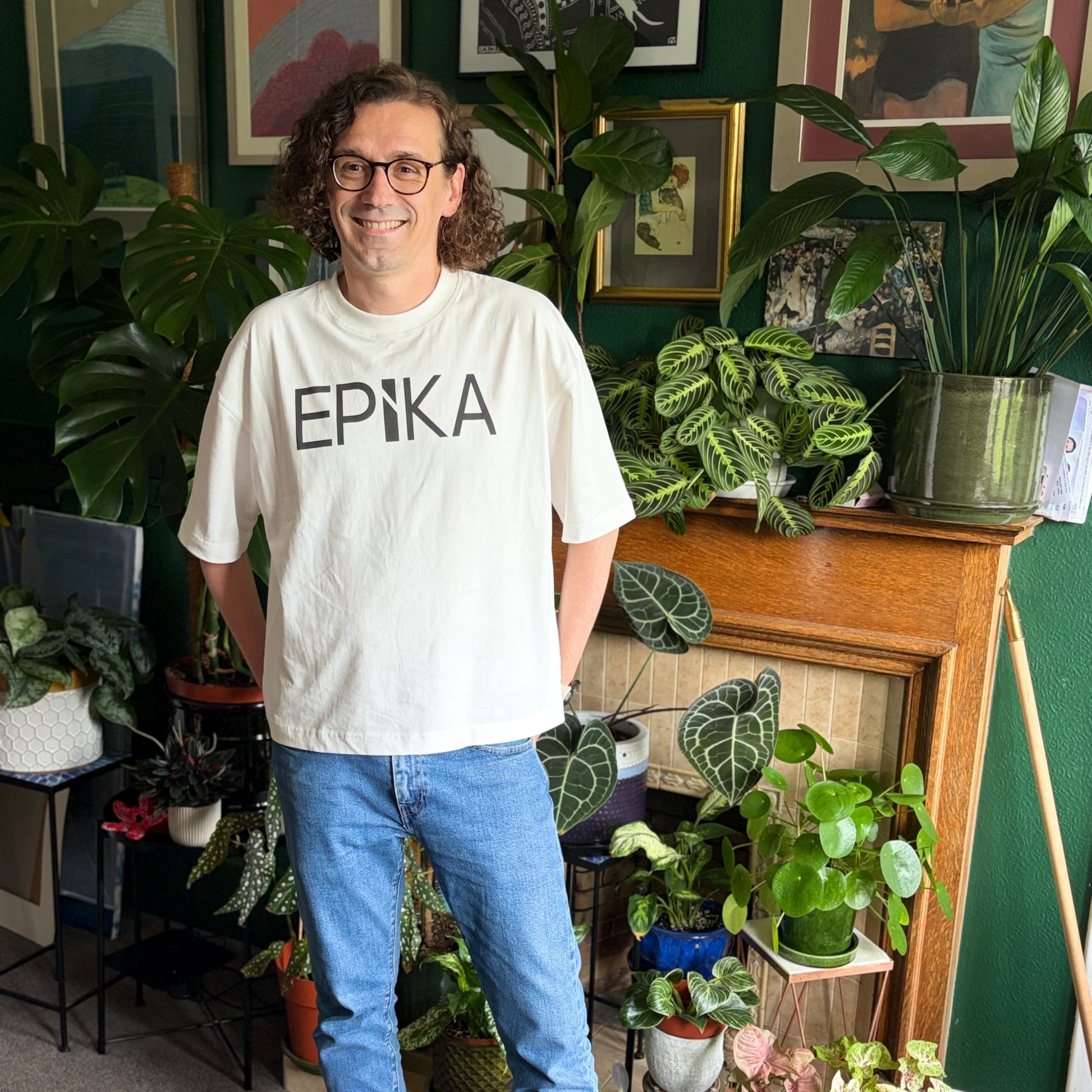 A man wearing glasses, a cream t-shirt with 'EPiKA' printed in black, blue jeans, and green sneakers, standing in a plant-filled room with framed art on the wall.